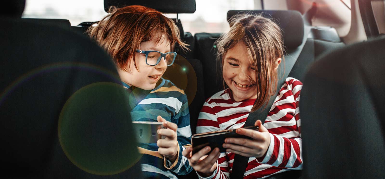 Two young kids in the backseat of a car looking at phones.